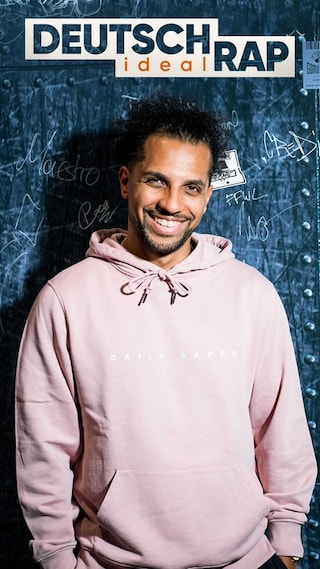 Deutschrap ideal-Host Simon sitzt seitlich mit dem Blick in die Kamera auf einem Tisch vor einer grauen Wand 